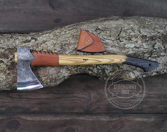 May include: A hand-forged axe with a brown leather handle wrap and a wooden shaft. The axe has a black stained handle and a polished steel head. The axe is resting on a wooden log with a leather sheath.