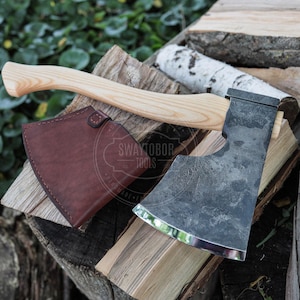 Puede incluir: Un hacha de mano con mango de madera clara y cabeza de metal oscuro texturizado con filo pulido. Una funda de cuero marrón con cierre de broche se sitúa junto a la cabeza del hacha. El texto SWAYTOBOR TOOLS es visible en la superficie.