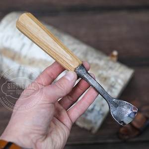 May include: A small, hand-crafted wood carving tool with a wooden handle and a curved metal blade. The tool is held in a person's hand.