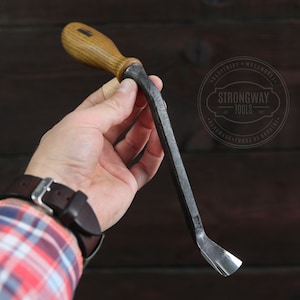 May include: A hand holds a small, curved woodworking tool with a wooden handle. The tool is made of metal and has a sharp, angled blade. The tool is labeled "Strongway Tools" with the text "Carpentry - Woodworking - Handmade - Forge of Darwin".