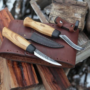 May include: Two wood-handled knives with leather sheaths. The knife on the left has a curved blade and the knife on the right has a straight blade. The knives are on a brown leather sheath with the text "SWAYTOBOR TOOLS" printed on it.