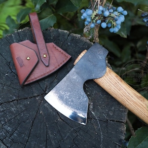 May include: A hand axe with a wooden handle and a steel blade, next to a brown leather sheath. The axe head has a textured finish. The sheath has a loop for carrying and is secured with brass rivets.