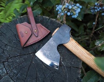 May include: A hand axe with a wooden handle and a steel blade, alongside a brown leather sheath. The axe head has a textured finish. The sheath has a loop for carrying and is secured with brass rivets.