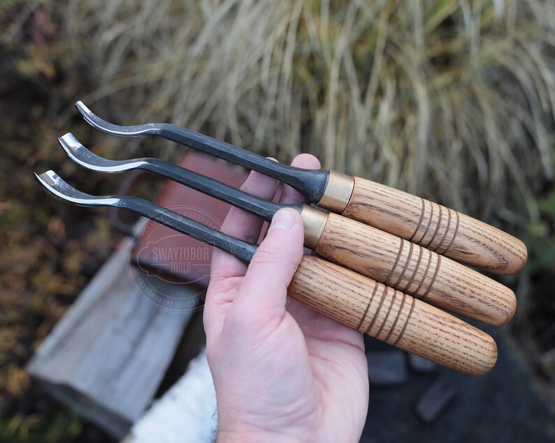 Może przedstawiać: Trzy zakrzywione narzędzia do rzeźbienia w drewnie z ciemnymi metalowymi ostrzami i drewnianymi uchwytami. Uchwyty są jasnobrązowe z ciemnobrązowymi pierścieniami i mosiężnymi tulejami. Rozmyte tło.