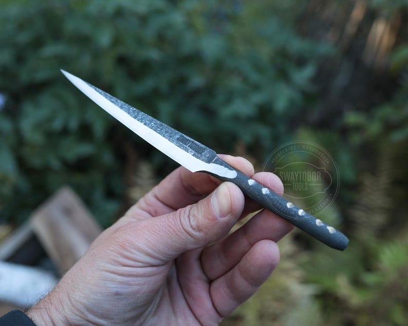 May include: A handmade knife with a silver blade and a dark gray handle. The blade is sharp and pointed, and the handle has a textured pattern with small, silver accents. The knife is held in a hand against a blurred green background.