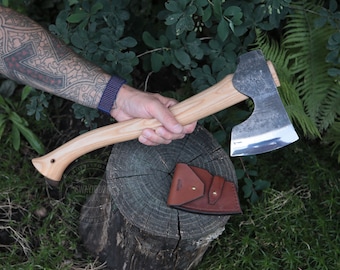 May include: A hand holds a wooden-handled axe with a metal head. The axe has a leather sheath with a metal snap. The axe is resting on a tree stump.