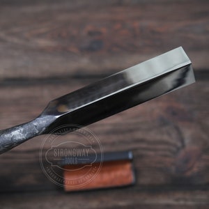 May include: A close-up of a black metal chisel with a beveled edge. The chisel is on a wooden surface with a brown leather case in the background. The case has the text "Strongway Tools" printed on it.