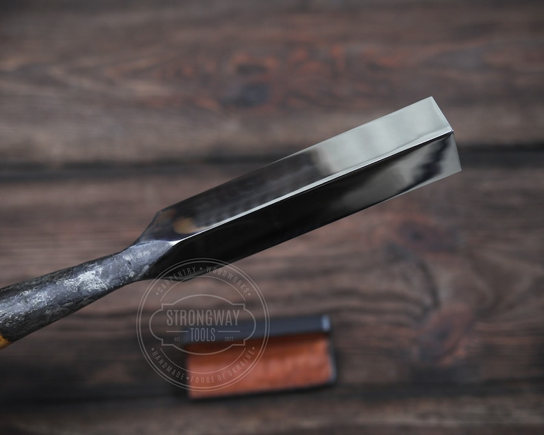 May include: A close-up of a black metal chisel with a beveled edge. The chisel is on a wooden surface with a brown leather case in the background. The case has the text "Strongway Tools" printed on it.
