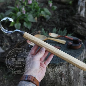 以下が含まれることがあります： 湾曲した刃を持つ木製のハンドル付きフックナイフ。ハンドルは明るい木で作られており、「Strongway Tools」という文字が刻まれています。ナイフはタトゥーのある手で持たれています。