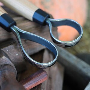 May include: Close-up of two carving tools with wooden handles and curved, polished metal blades. The tools have a black collar where the handle meets the blade. The background is blurred, suggesting an outdoor setting.