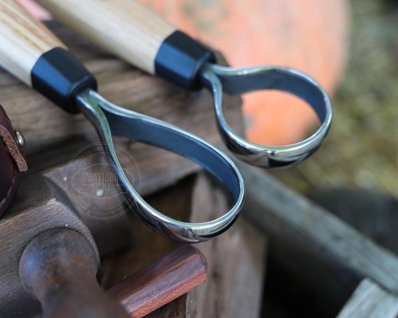 May include: Close-up of two carving tools with wooden handles and curved, polished metal blades. The tools have a black collar where the handle meets the blade. The background is blurred, suggesting an outdoor setting.