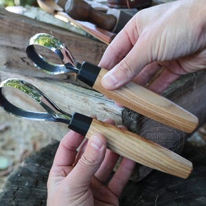 Puede incluir: Dos herramientas para tallar madera con hojas curvas de acero pulido y mangos de madera. Los mangos son de color marrón claro con un collar negro donde se encuentran con la hoja. Las herramientas se sostienen en las manos de una persona.