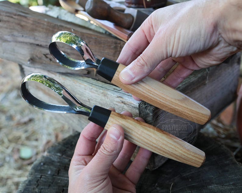 May include: Two wood carving tools with curved, polished steel blades and wooden handles. The handles are light brown with a black collar where they meet the blade. The tools are held in a person's hands.