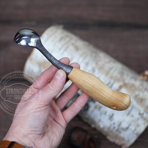 May include: A small, hand-forged spoon with a wooden handle. The spoon is made of metal and has a rounded bowl. The handle is made of wood and has a natural finish. The spoon is labeled "Strongway Tools" and "Handmade".