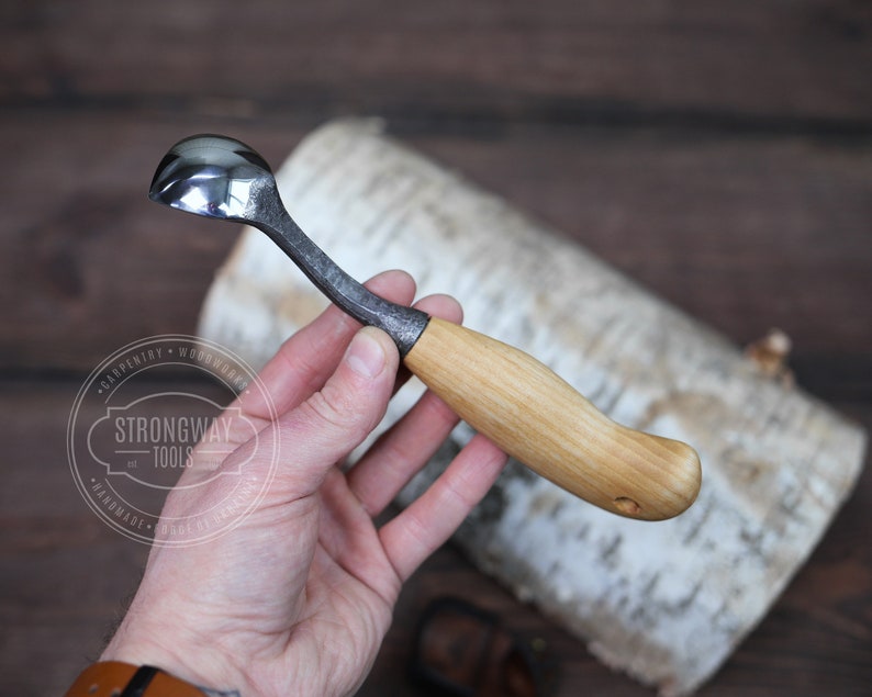 May include: A small, hand-forged spoon with a wooden handle. The spoon is made of metal and has a rounded bowl. The handle is made of wood and has a natural finish. The spoon is labeled "Strongway Tools" and "Handmade".