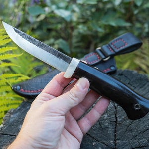 May include: A handcrafted knife with a dark wooden handle and a silver blade. The knife is held in a hand, with a black leather sheath visible in the background. The blade has a slightly textured appearance.