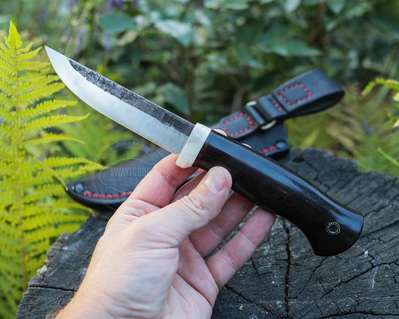 May include: A handcrafted knife with a dark wooden handle and a silver blade. The knife is held in a hand, with a black leather sheath visible in the background. The blade has a slightly textured appearance.