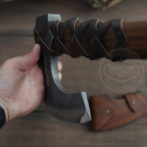May include: A hand holds a black and silver axe with a brown leather handle. The axe has a curved blade and a leather wrap around the handle. The axe is made by Strongway Tools.