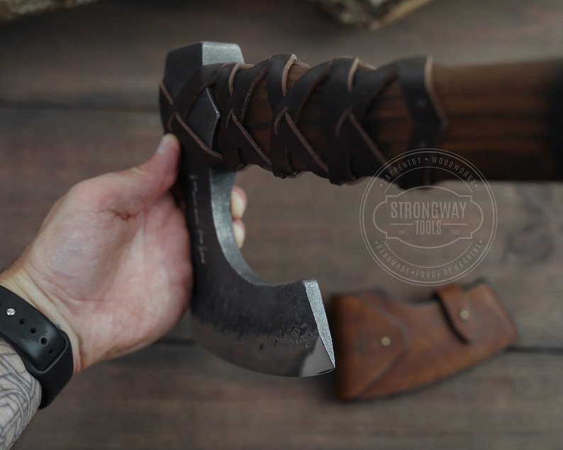 May include: A hand holds a black and silver axe with a brown leather handle. The axe has a curved blade and a leather wrap around the handle. The axe is made by Strongway Tools.