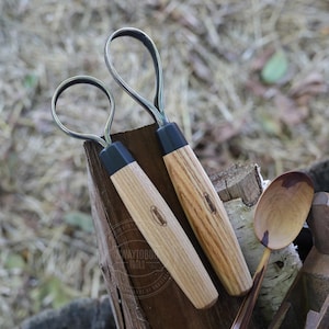 May include: Two wood carving tools with curved metal blades and wooden handles. The handles are light brown with a black base. The tools are resting on a wooden surface with a wooden spoon and other woodworking tools.