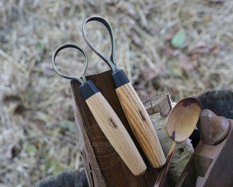 May include: Two wood carving tools with curved metal blades and wooden handles. The handles are light brown with a black base. The tools are resting on a wooden surface with a wooden spoon and other woodworking tools.