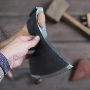 May include: A hand holds a large, black, single-bevel axe with a wooden handle. The axe has a curved blade and a rounded head. The axe is on a wooden surface.