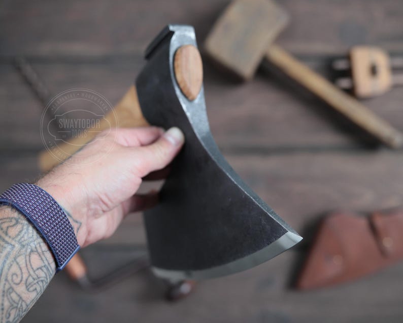 May include: A hand holds a large, black, single-bevel axe with a wooden handle. The axe has a curved blade and a rounded head. The axe is on a wooden surface.