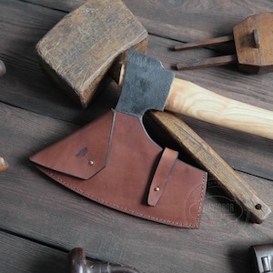 May include: A brown leather axe sheath with a black metal axe head and a wooden handle. The sheath has a stitched edge and a metal rivet.