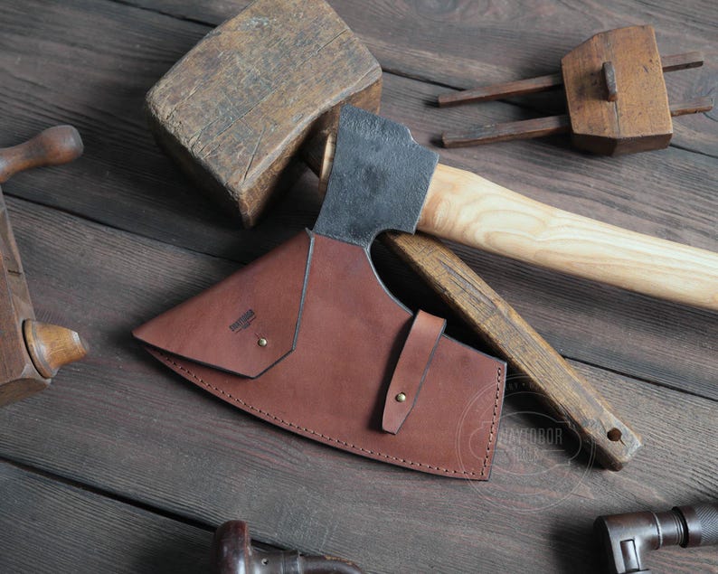 May include: A brown leather axe sheath with a black metal axe head and a wooden handle. The sheath has a stitched edge and a metal rivet.