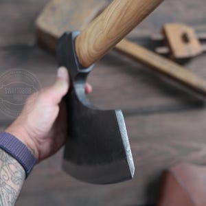 May include: A hand holds a black axe with a wooden handle. The axe has a curved blade and a sharp edge. The axe is on a wooden surface.