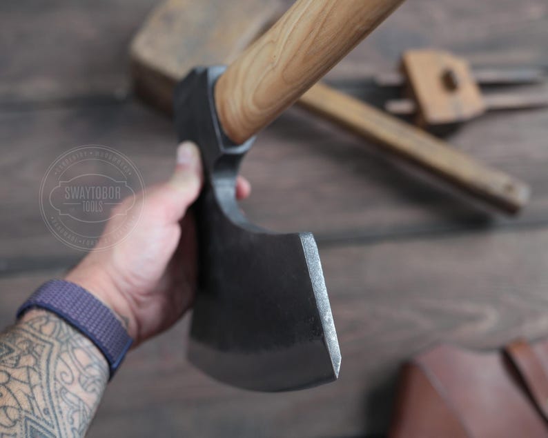 May include: A hand holds a black axe with a wooden handle. The axe has a curved blade and a sharp edge. The axe is on a wooden surface.