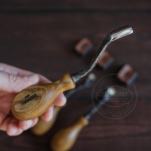 May include: A close-up of a wood carving tool with a curved blade and a wooden handle. The handle is engraved with the words "Strongway Tools".