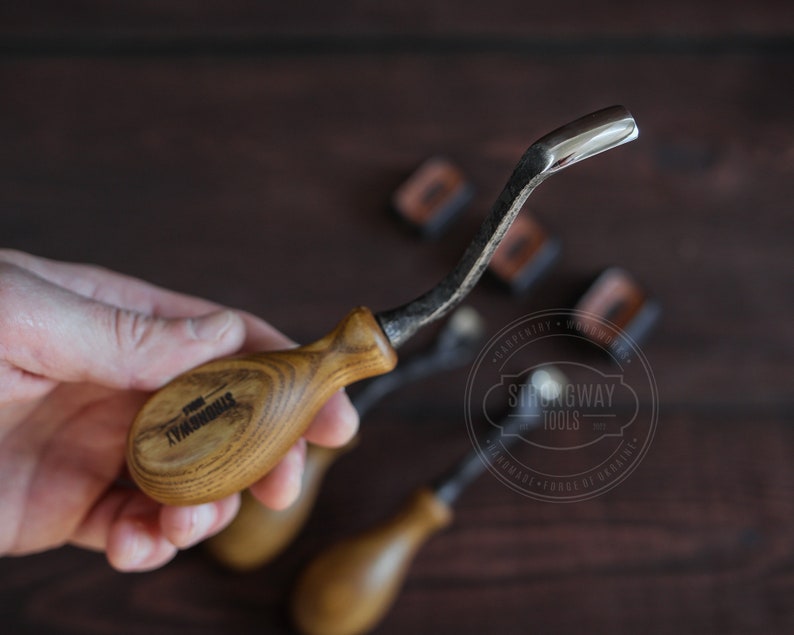 May include: A close-up of a wood carving tool with a curved blade and a wooden handle. The handle is engraved with the words "Strongway Tools".