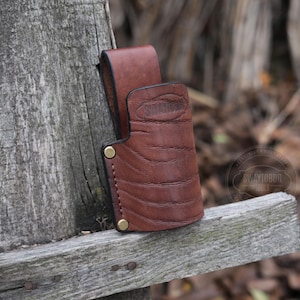 May include: A brown leather sheath with brass rivets and the word "SWAYTOBOR" embossed on the front. The sheath is hanging on a wooden fence.