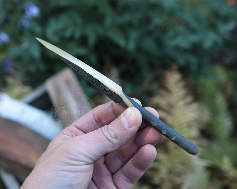 May include: A handcrafted, small, dark-colored wooden knife with a light-colored blade. The knife is held in a person's hand against a blurred green background. The blade appears to be approximately 4 inches long.