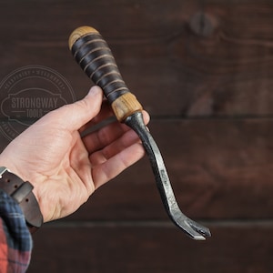 May include: A hand holds a curved, metal nail puller with a wooden handle. The handle is wrapped in brown leather and has a dark brown stain. The tool is called a "Strongway Tools" nail puller.