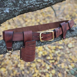 Puede incluir: Un cinturón de cuero marrón con una hebilla de latón y múltiples bolsas de cuero adjuntas. El cinturón está colgado de una rama de árbol.