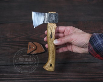 May include: A small, hand-crafted axe with a wooden handle. The axe head is made of steel and has a polished finish. The handle is made of wood and has a natural finish. The axe is being held by a hand in the image. The axe is labeled "Strongway Tools".