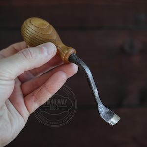 May include: A small, hand-forged wood carving tool with a curved blade and a wooden handle. The tool is held in a hand, and the background is a dark wood surface. The tool is labeled "Strongway Tools est. 2022 Handmade - Forge of Ukraine."