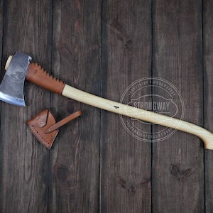 Könnte beinhalten: Eine handgeschmiedete Axt mit einem Ledergriff und einer braunen Lederscheide. Die Axt liegt auf einem Holzgrund. Der Text "Strongway Tools" ist auf dem Griff sichtbar.