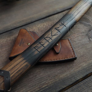 May include: A wooden-handled axe with a black finish and a leather sheath. The handle is engraved with runes and the text "Strongway Tools 1990".