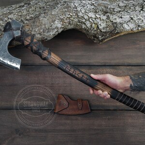 May include: A hand holds a black and brown axe with a leather handle. The axe head is silver and has a hammered finish. The axe handle is engraved with the word "Viking".