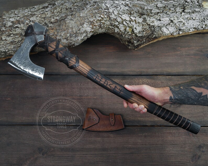 May include: A hand holds a black and brown axe with a leather handle. The axe head is silver and has a hammered finish. The axe handle is engraved with the word "Viking".