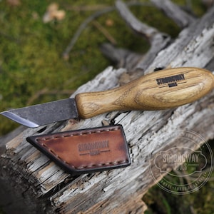 Puede incluir: Un pequeño cuchillo de talla de madera, forjado a mano, con mango de madera y funda de cuero. El cuchillo tiene una hoja afilada y puntiaguda y está grabado con las palabras "Strongway Tools".