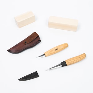 Könnte beinhalten: Zwei Holzschnitzmesser mit Holzgriffen und scharfen Klingen, zusammen mit einer braunen Lederscheide und zwei Holzblöcken. Die Messer haben eine helle Holzoberfläche und sind für detaillierte Schnitzereien konzipiert.