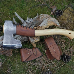 Op de afbeelding: Een handgesmeed bijl met een bruin leren handvat en een houten steel. De bijl ligt op een stuk hout met groen mos. De bijl wordt geleverd met een bruine leren schede.