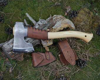 May include: A hand-forged axe with a brown leather handle and a wooden shaft. The axe is resting on a piece of wood with green moss. The axe comes with a brown leather sheath.