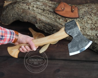 May include: A hand holds a black and silver axe with a wooden handle. The axe has a slightly curved blade and a textured surface. The handle is made of light wood and has a slight curve at the end. The axe is resting on a wooden surface.