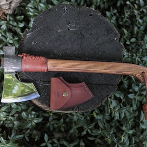 Könnte beinhalten: Ein Handbeil mit poliertem Metallkopf, Holzstiel und braunem Lederband nahe dem Kopf. Eine braune Lederscheide mit Messingdruckknopf befindet sich unter dem Axtkopf. Eine dünne Lederschnur ist am Ende des Stiels befestigt.