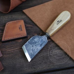 Puede incluir: Una pequeña herramienta de trabajo del cuero forjada a mano con un mango de madera. La herramienta tiene una cuchilla de metal y está etiquetada como "Strongway Tools".
